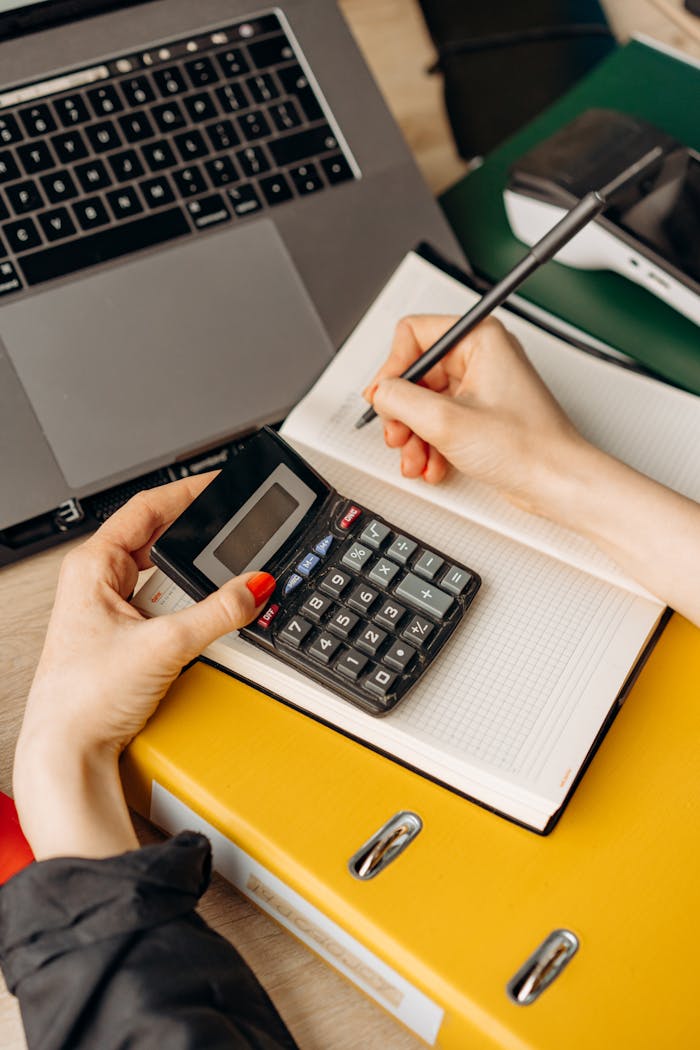 Hands using a calculator and writing on a notebook at a modern workspace with laptop.